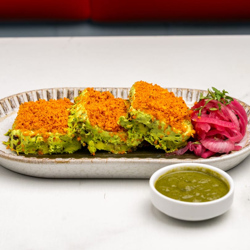Three green Indian dishes topped with orange crumbs, served with pickled onions and green chutney.