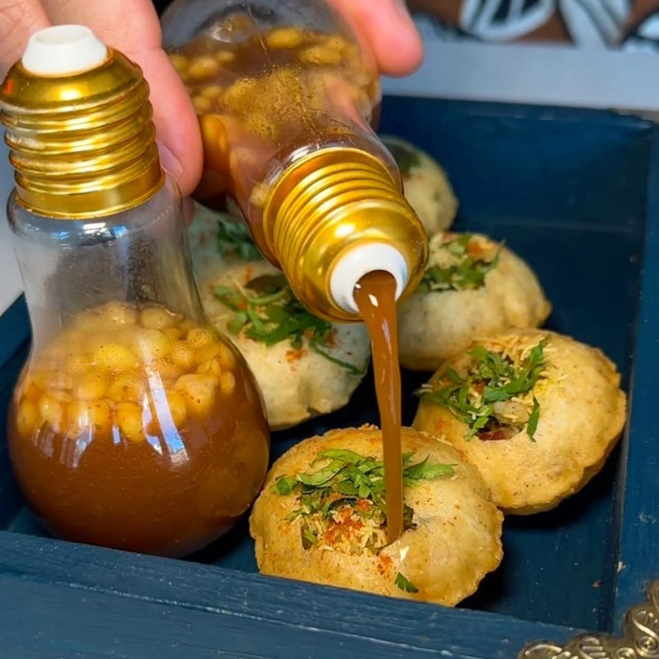 A hand pours tamarind water from a lightbulb-shaped bottle into a pani puri on a blue tray.