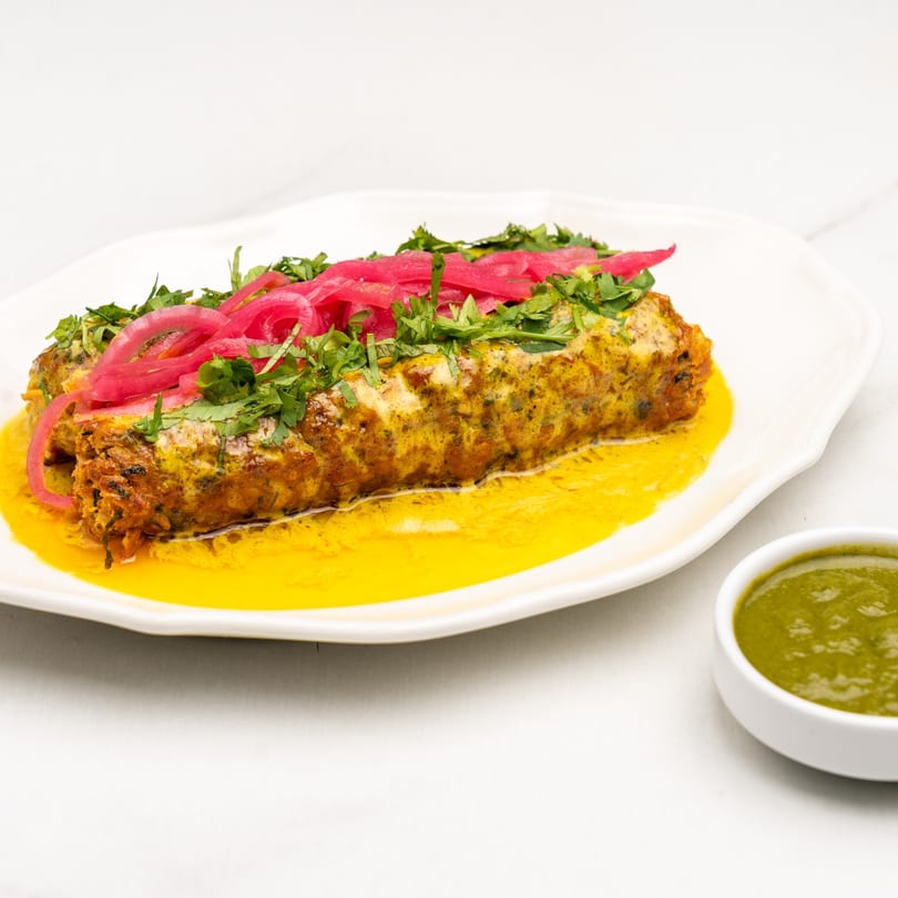 Indian kebab on a white plate, garnished with pickled red onions and fresh cilantro, with a side of green chutney.